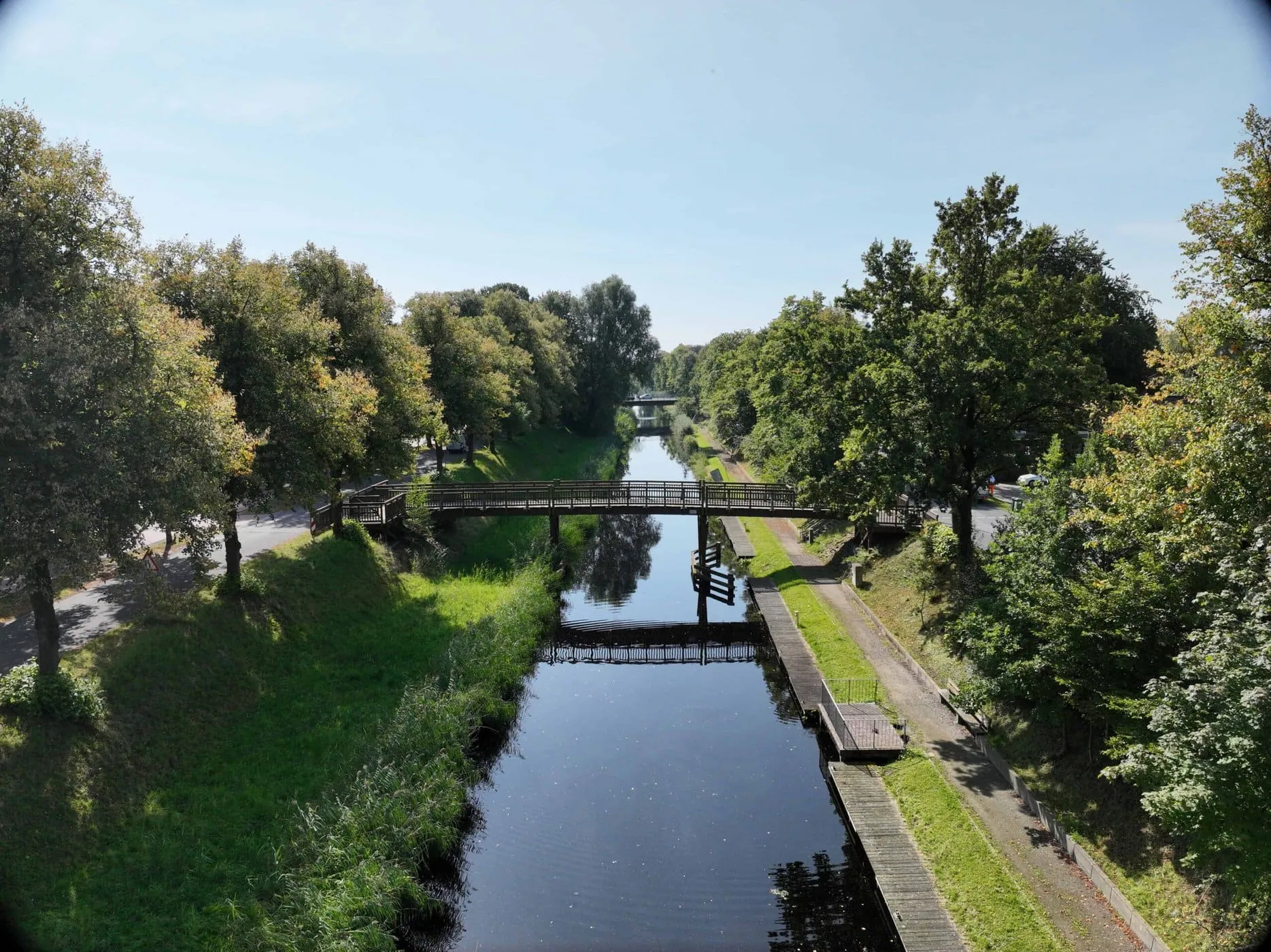 Ein malerischer Kanal mit einer hölzernen Fußgängerbrücke und grünen Bäumen auf beiden Seiten unter einem klaren blauen Himmel.